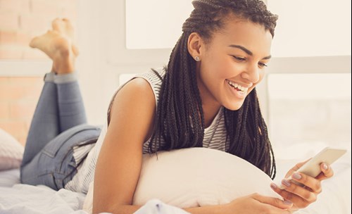 Young woman lying on bed, smiling while busy on smartphone