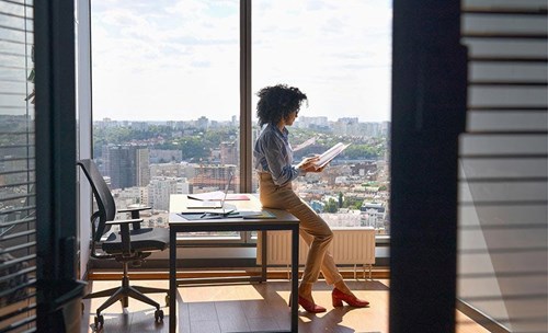 Woman sitting on desk in a office, with city view in the background