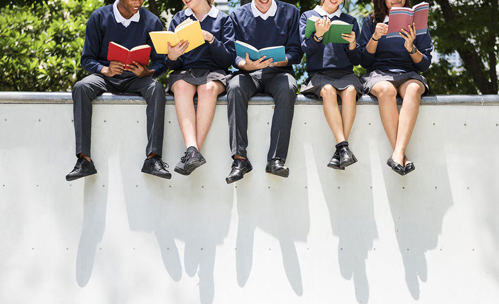 Children in school uniform sitting on a wall, reading books, with their legs hanging over the wall