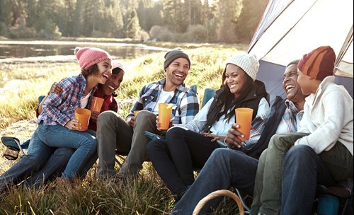 Laughing group of people sitting on camping chairs next to a tent in nature, water feature and trees in the background