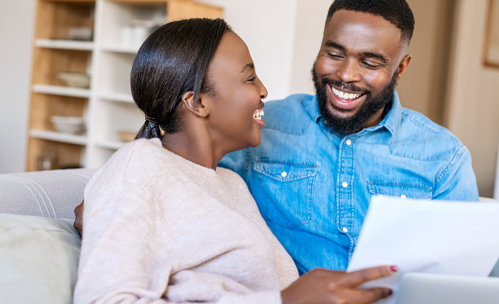 Smiling couple sitting on couch, looking at each other, while holding documents