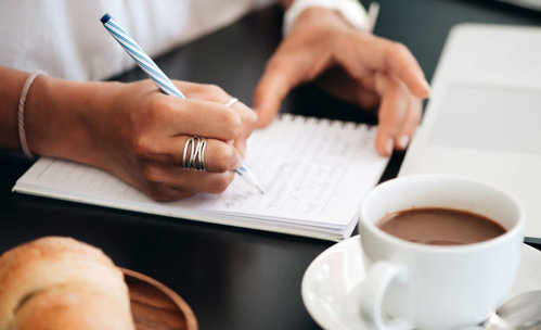 Woman writing in a book, with laptop and coffee on the table